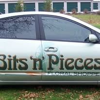 Side of a car with Bits n' Pieces Floral Shoppe logo. The car is light-colored and parked near buildings with garage doors.