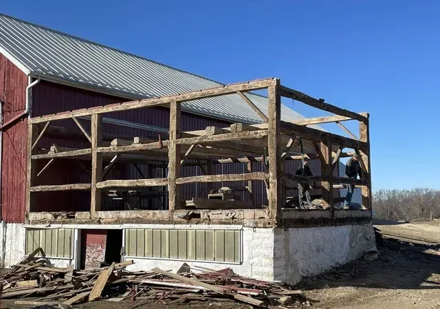 Barn with exposed wooden frame under repair; red exterior, white foundation, blue sky.