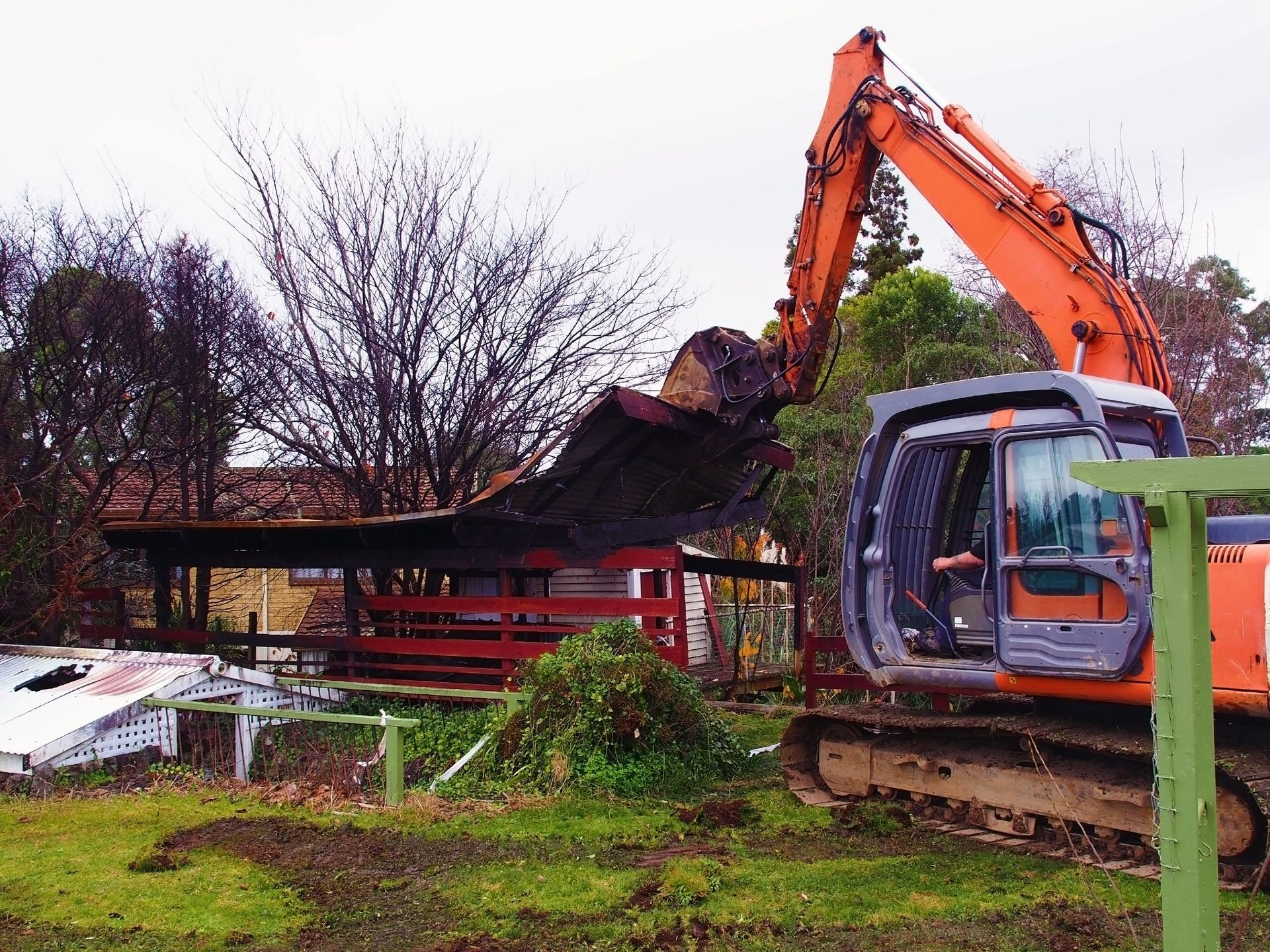 Orange excavator demolishing a shed in a yard on an overcast day.