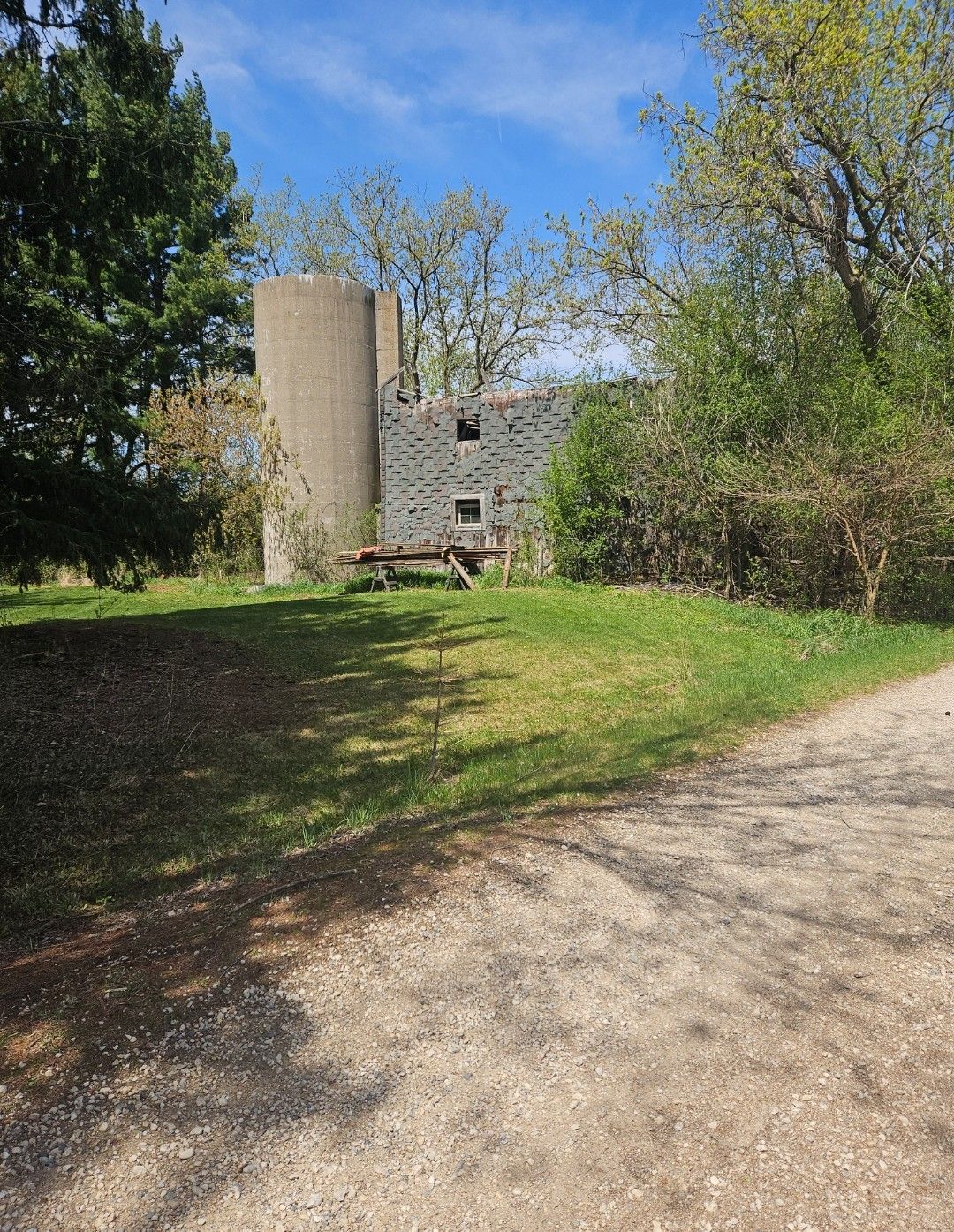 Ruins of a stone building with a cylindrical silo, surrounded by trees and grass, on a sunny day.