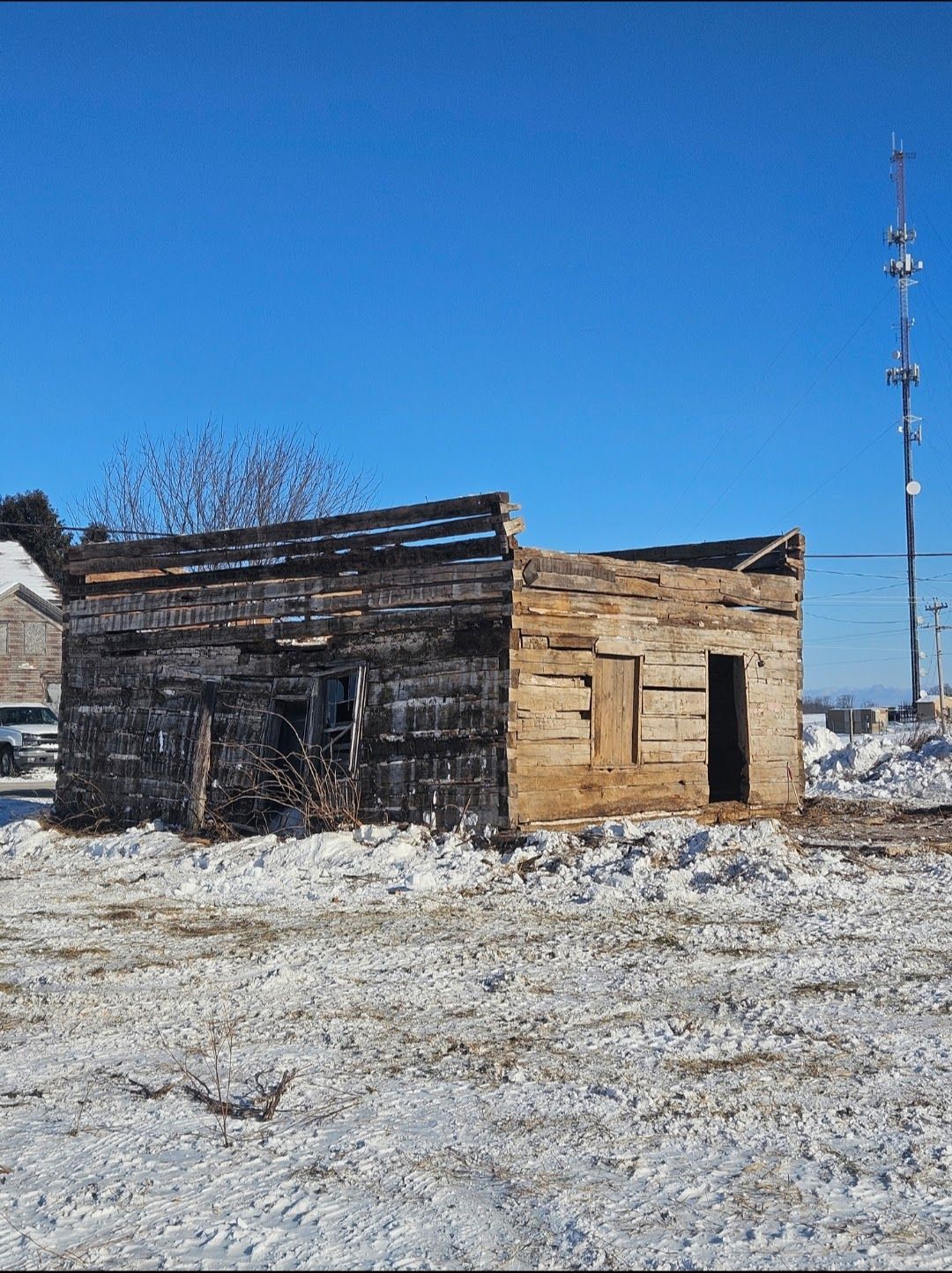 Dilapidated wooden building, possibly a cabin, in a snow-covered field under a clear, blue sky.
