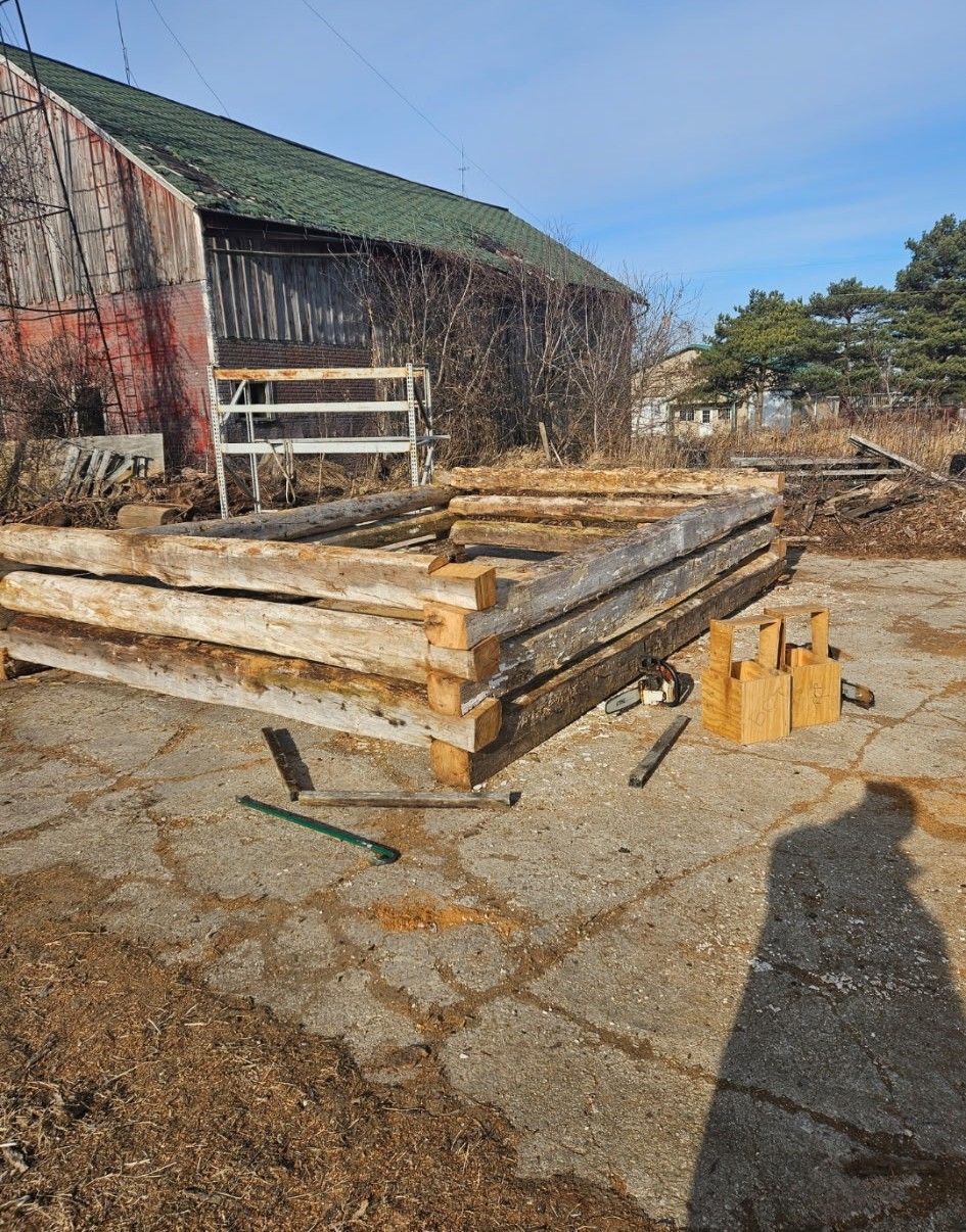 A rectangular raised garden bed made of stacked wooden logs sits on a concrete pad near a rustic barn.