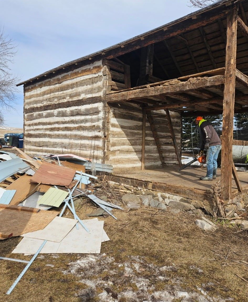 A person uses a chainsaw to dismantle a rustic wooden log structure with an open-sided porch amid a pile of debris.