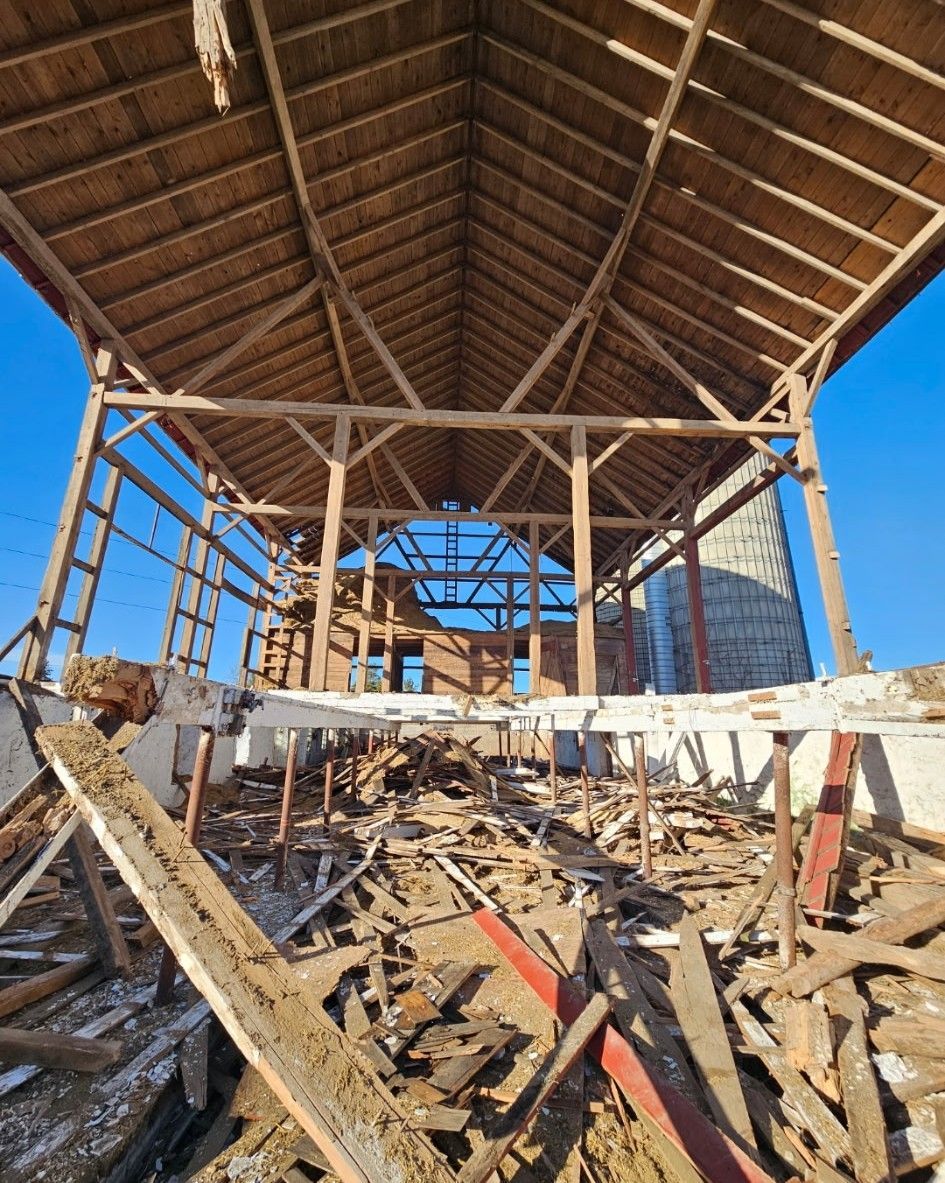 A wide-angle, low-view of a dilapidated barn with exposed wooden rafters, scattered debris, and a silo in the background.