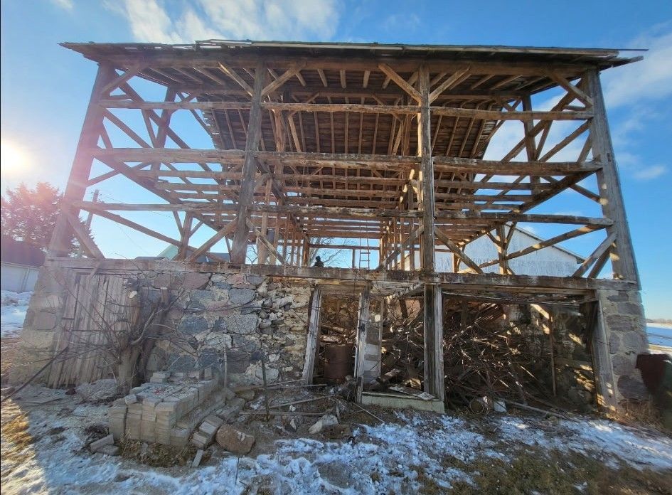 A weathered, open-frame barn structure stands on a stone foundation against a bright, winter sky with snow on the ground.