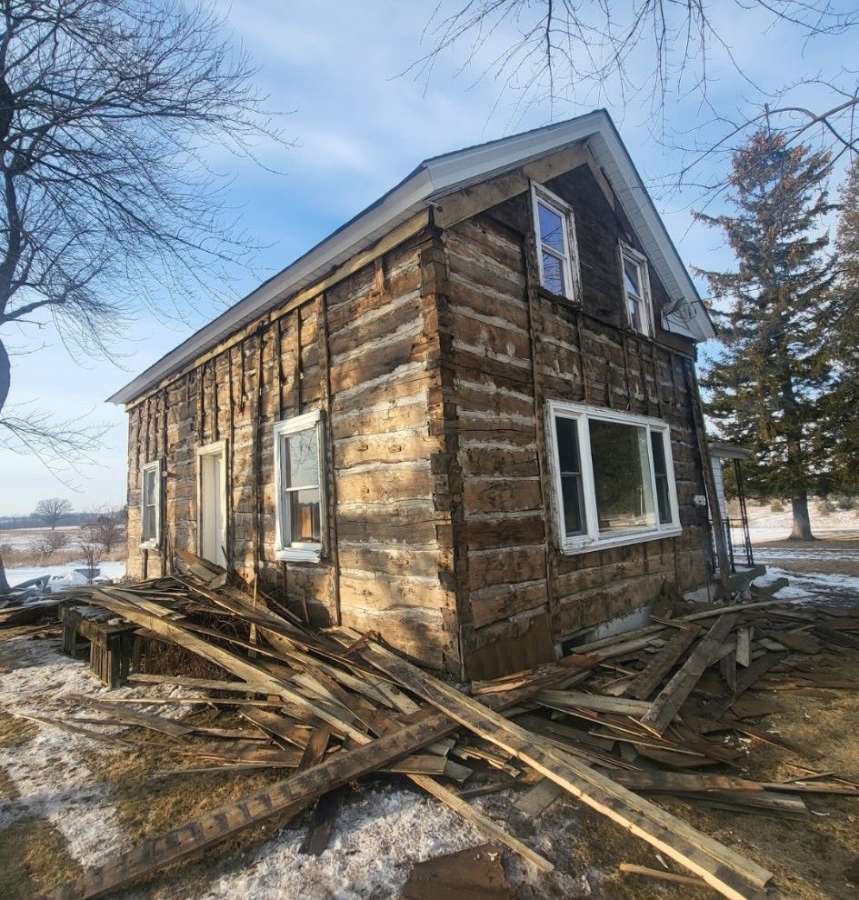 An old log cabin undergoing exterior siding removal, with discarded wooden boards piled on the ground outside.