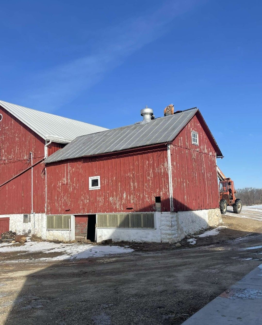 Red barn with peeling paint and metal roof against a clear blue sky. A tractor sits nearby.