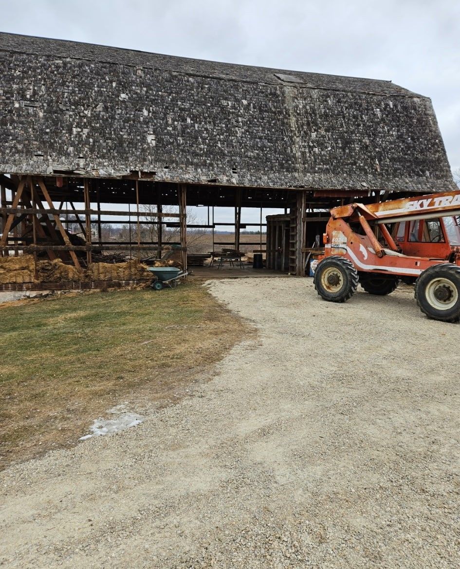 A weathered, open-sided wooden barn with a dark roof, next to a gravel path and an orange telehandler.