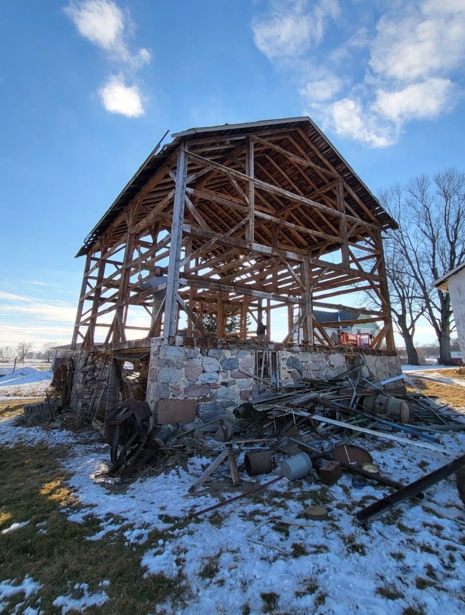 A skeletal wooden barn frame stands atop a stone foundation in a snowy, sunlit field under a blue sky.