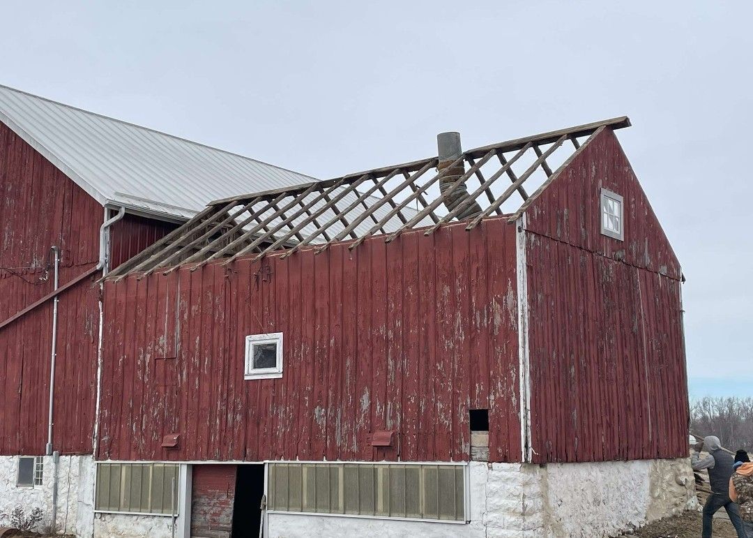 Red barn with missing roof section, revealing rafters; weathered wood, cloudy sky.