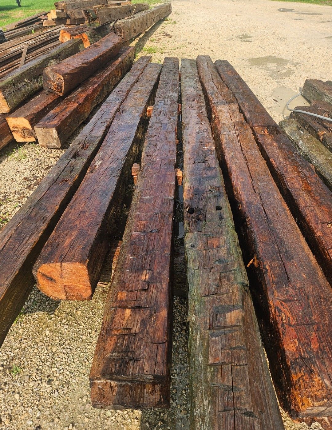 Pile of weathered, dark brown wooden beams, possibly railroad ties, outdoors on gravel.