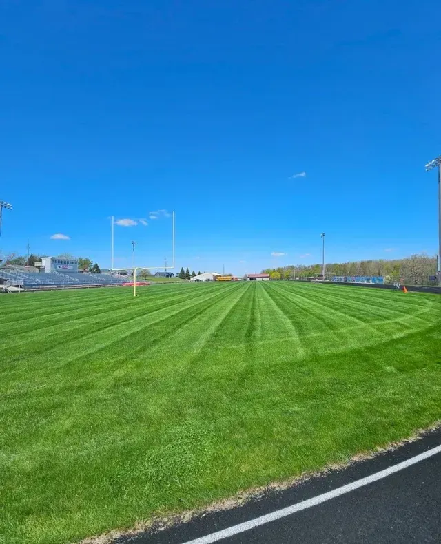A freshly mowed, striped football field viewed from the track under a clear blue sky.
