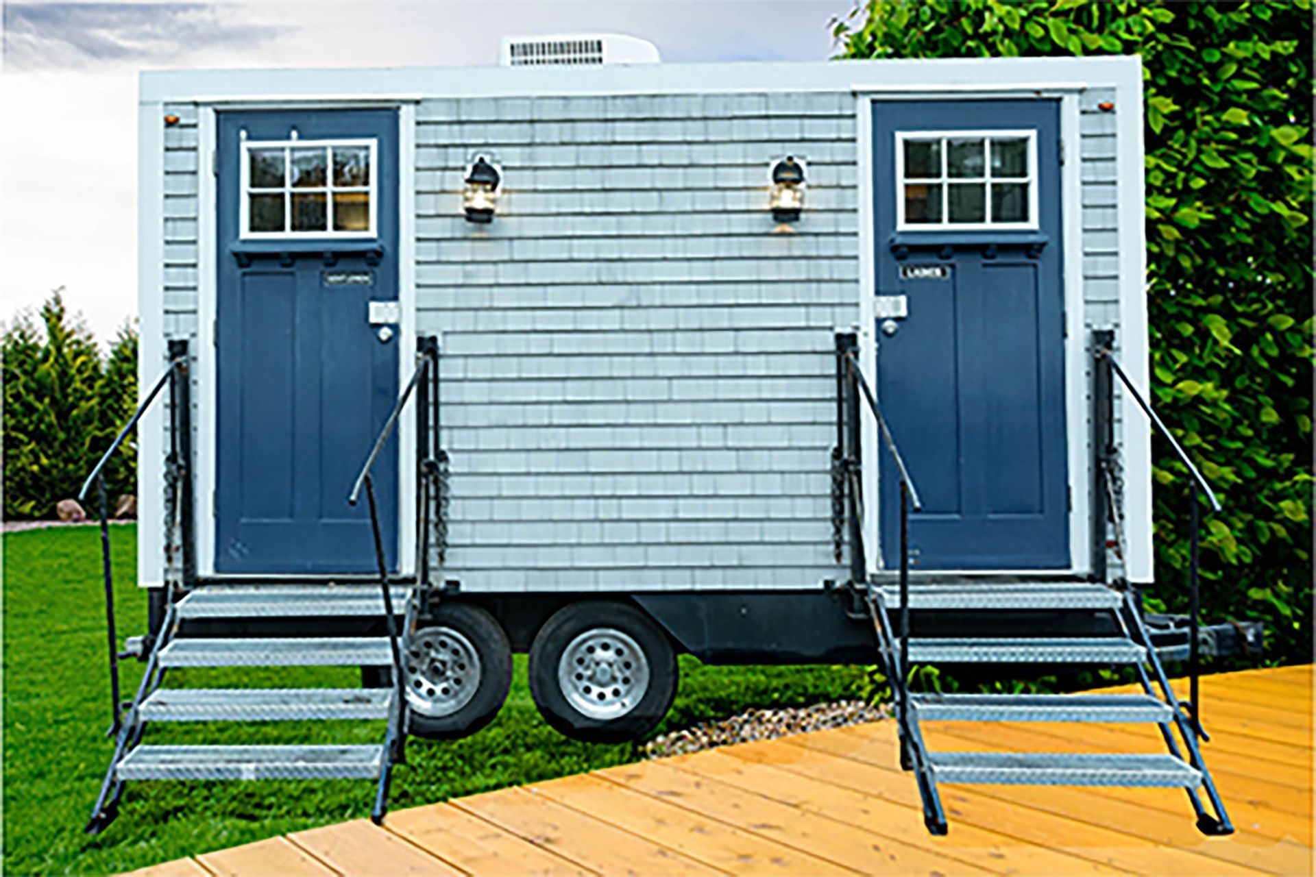 Blue tiny house trailer with two doors and steps on a wooden deck by grass and trees