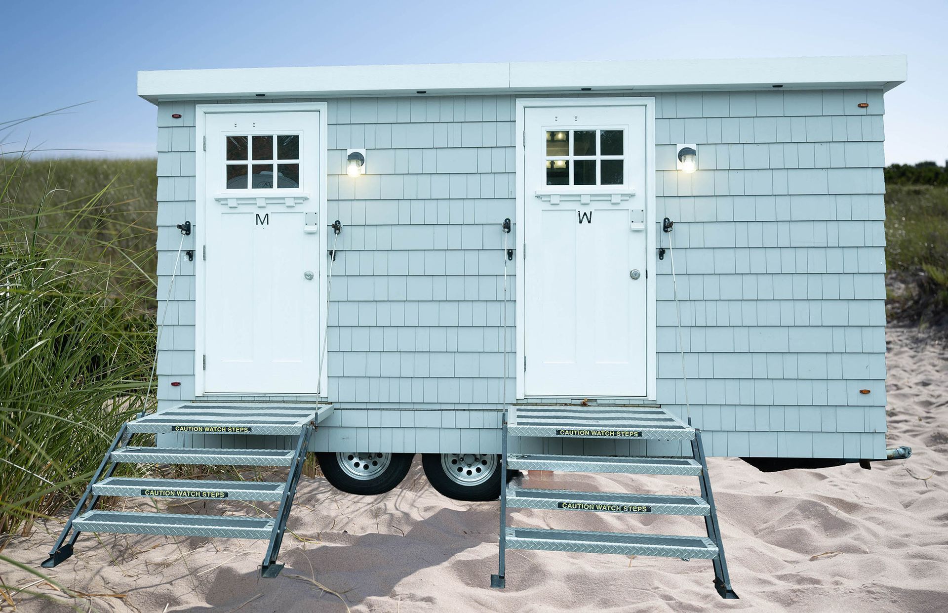 Two white beach huts with steps on sand, each with a door and small window, under a cloudy sky