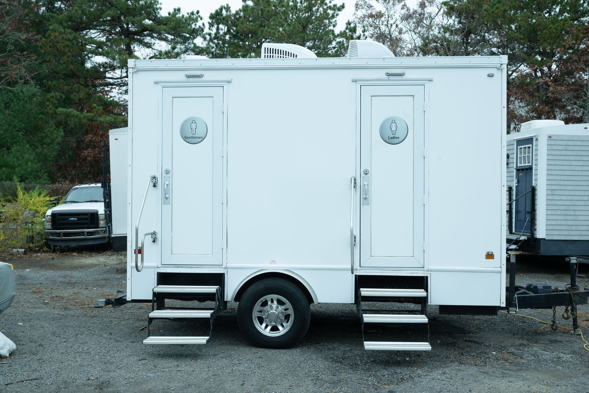 White portable restroom trailer with two doors, steps, and a truck in the background.