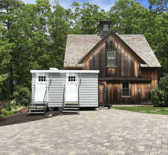 Wooden barn-style building with two-door, gray outhouse in front, surrounded by trees.