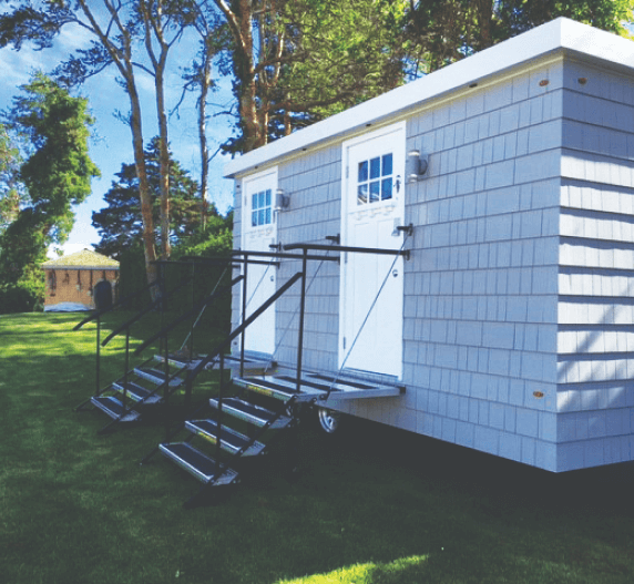 Gray, shingled, two-stall restroom building with stairs and handrails, set on green lawn.