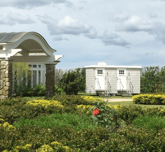 Stone structure with archway and windows, lawn with shrubs, and two small white outhouses with steps.