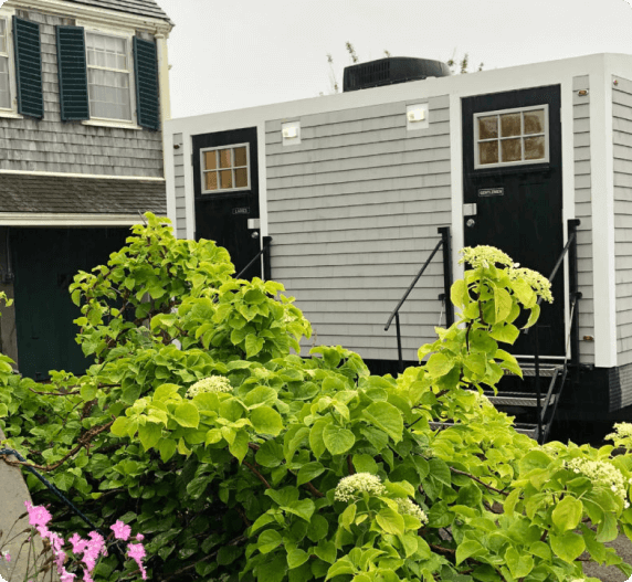 A luxury portable restroom trailer, light gray siding, black doors, small windows, green foliage.