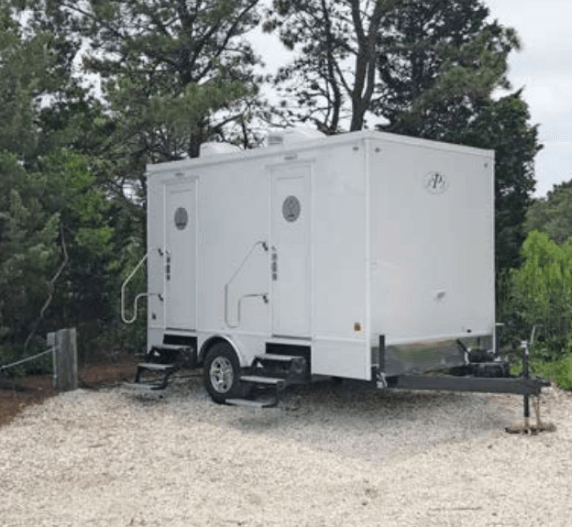 White portable restroom trailer on a gravel surface, surrounded by trees.