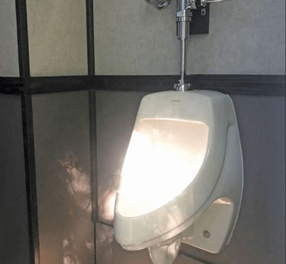 White urinal in a public restroom, with bright light illuminating its interior. Gray walls and plumbing visible.