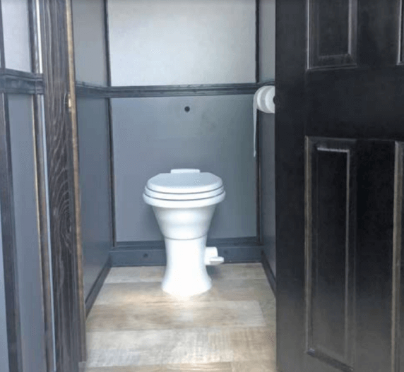 Interior of a portable toilet with a white toilet, gray walls, and a roll of toilet paper.