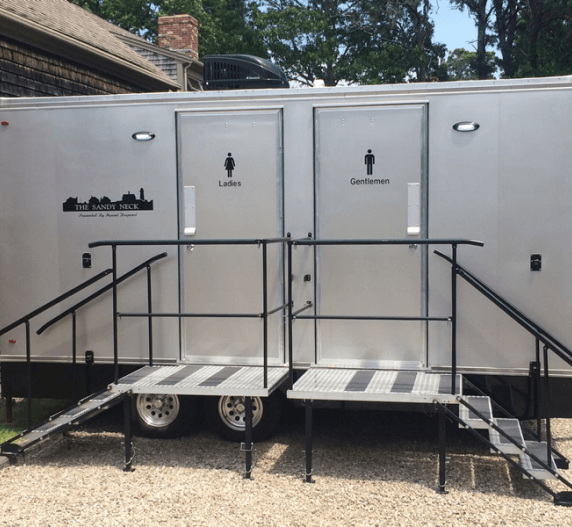 Portable restroom trailer with ramps, two doors labeled “Ladies” and “Gentlemen.”