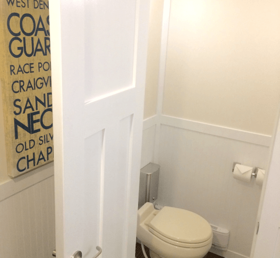 White-paneled powder room with toilet. Ajar white door reveals a Coast Guard sign. Neutral walls, wooden floor.