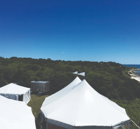 White tents atop a green hillside, ocean in the distance under a blue sky.