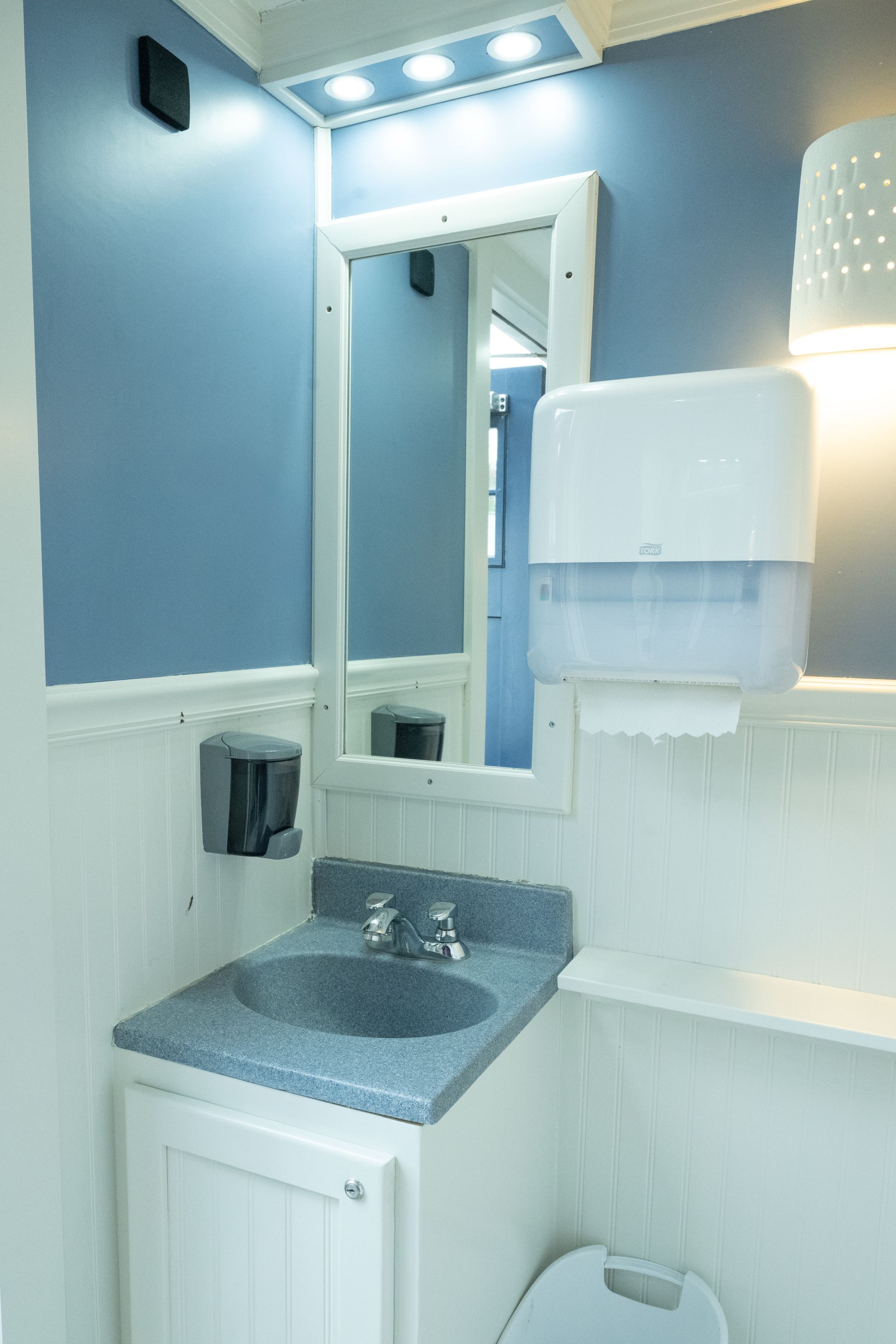 Interior of a portable restroom with a sink, mirror, and paper towel dispenser. Blue and white color scheme.