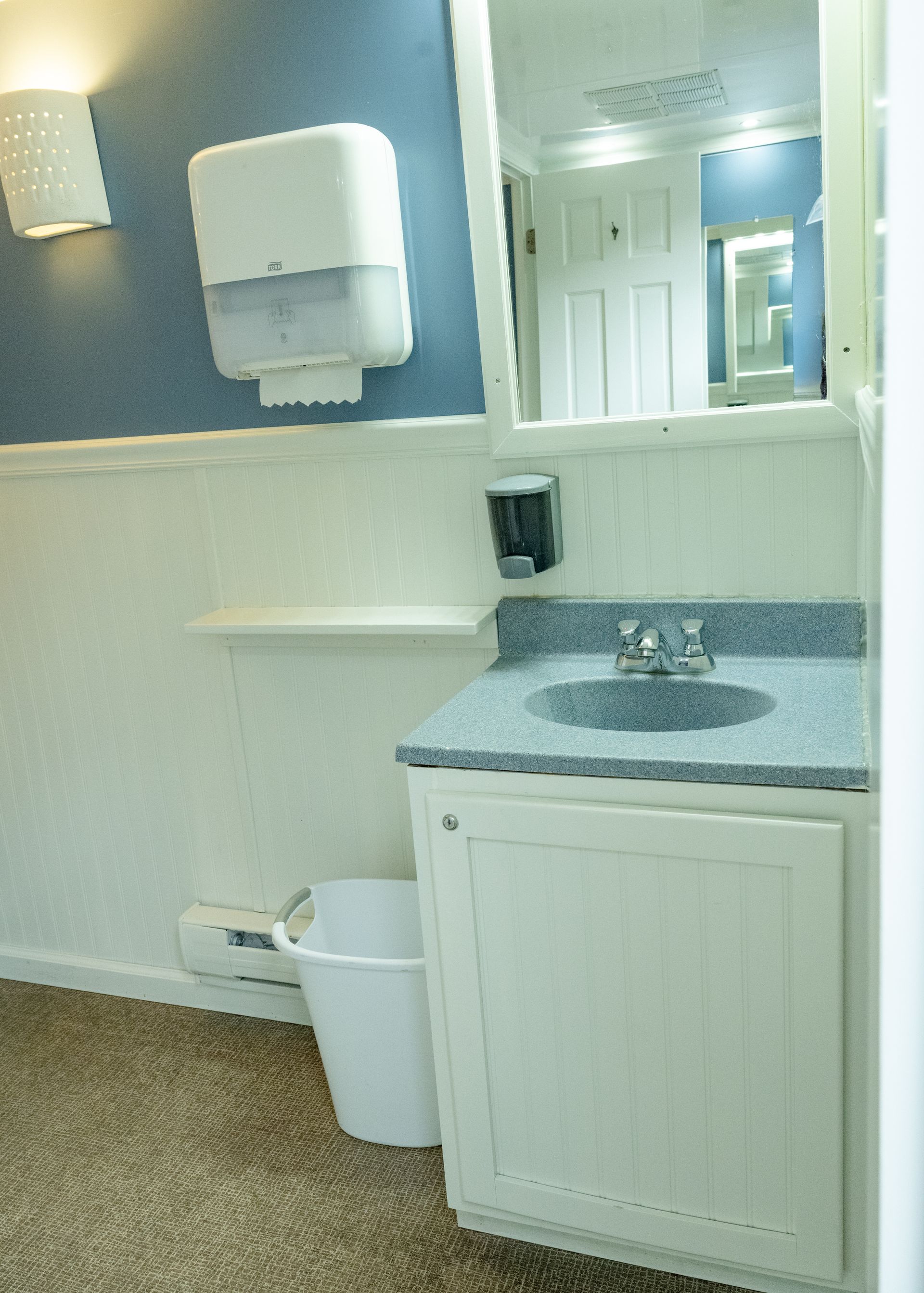 Interior of a portable restroom with a sink, mirror, and paper towel dispenser. White walls, blue wall above wainscoting.
