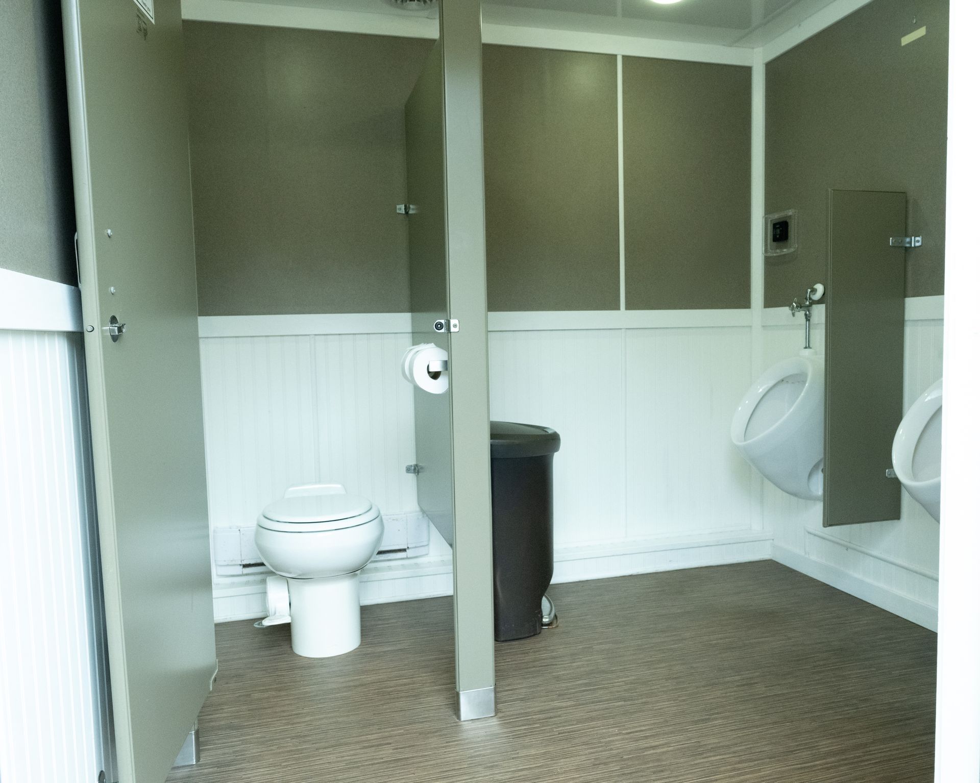 Interior of a portable restroom with a toilet, urinals, and trash can. Light gray walls and flooring.