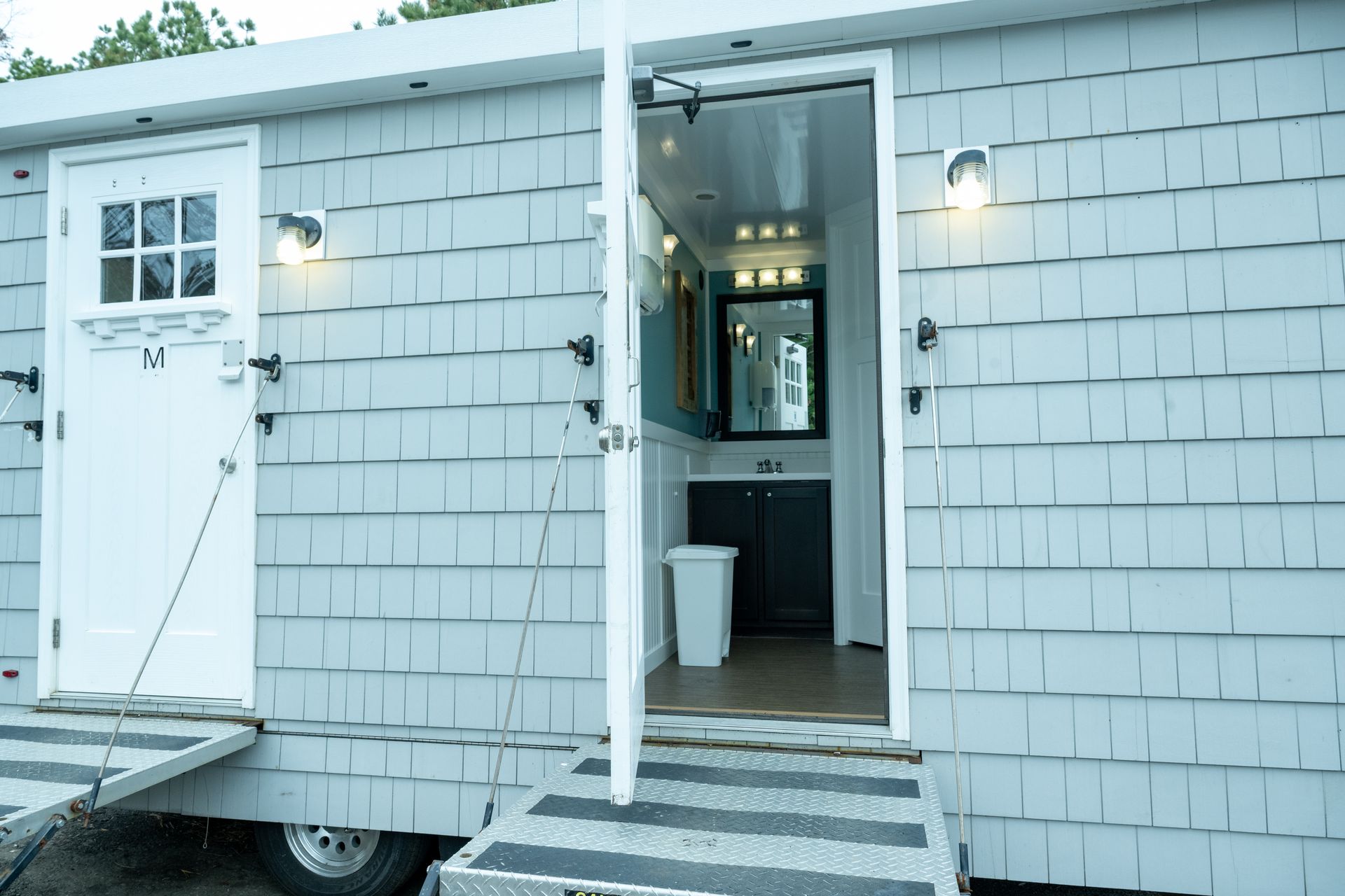 Portable restroom trailer with open door, gray siding, wheelchair ramp, and lights.
