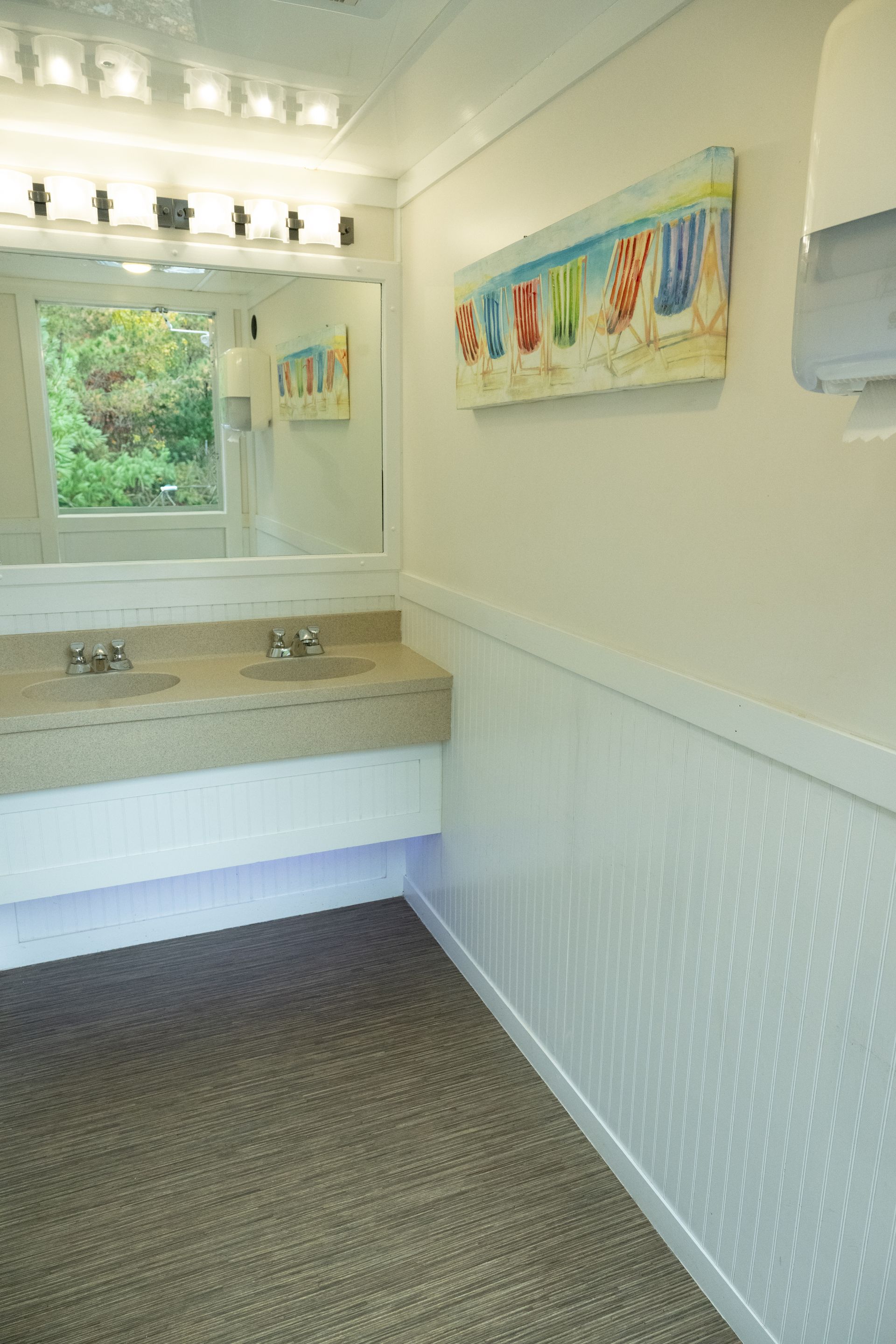 Interior of a restroom with a sink, mirror, and artwork of flip-flops. White walls and carpeted floor.