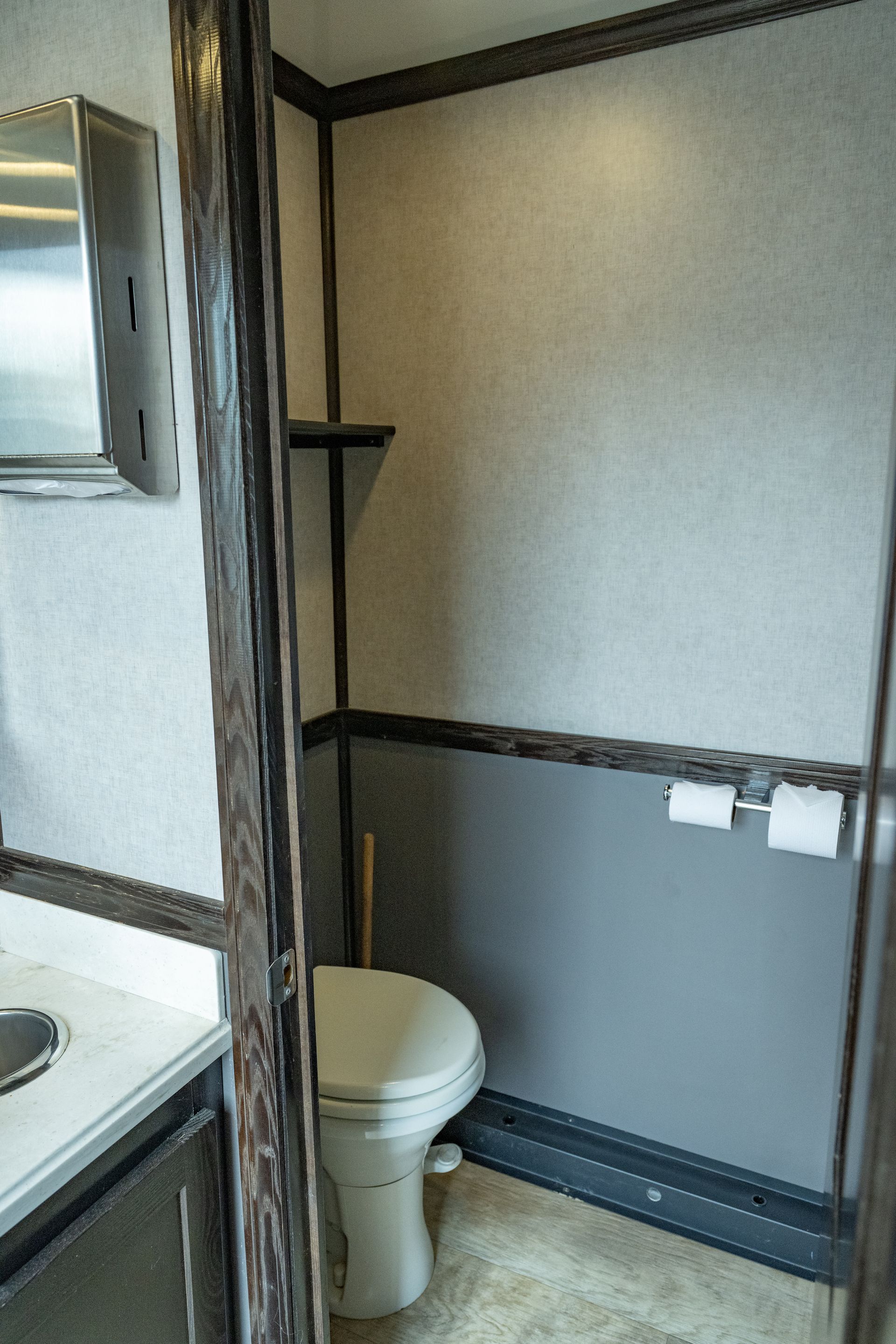 Interior of a portable restroom with a toilet, shelf, and a sink. Gray and white walls, dark trim.