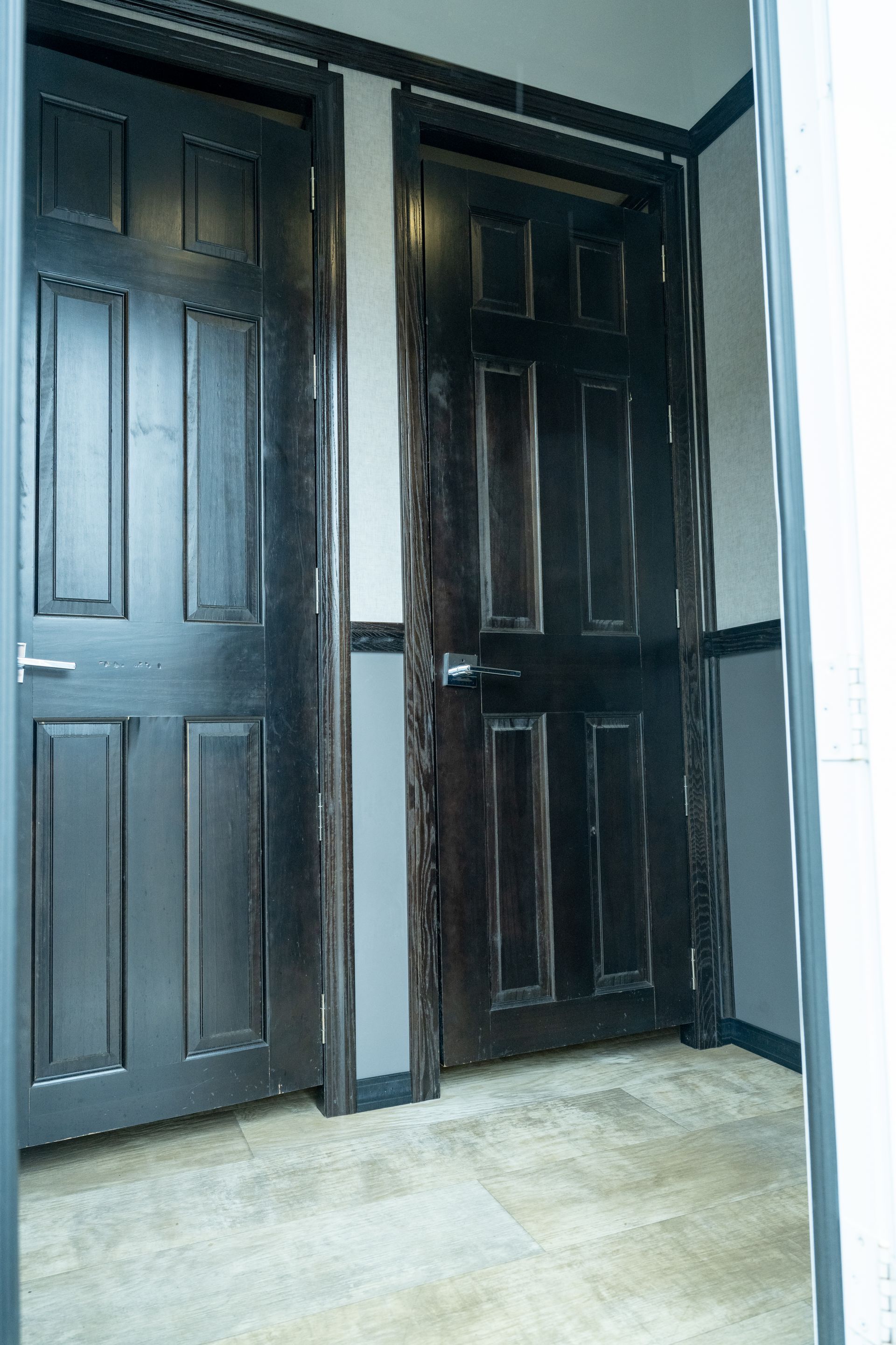 Two dark wooden doors, one open, in a hallway with light-colored flooring and gray walls.