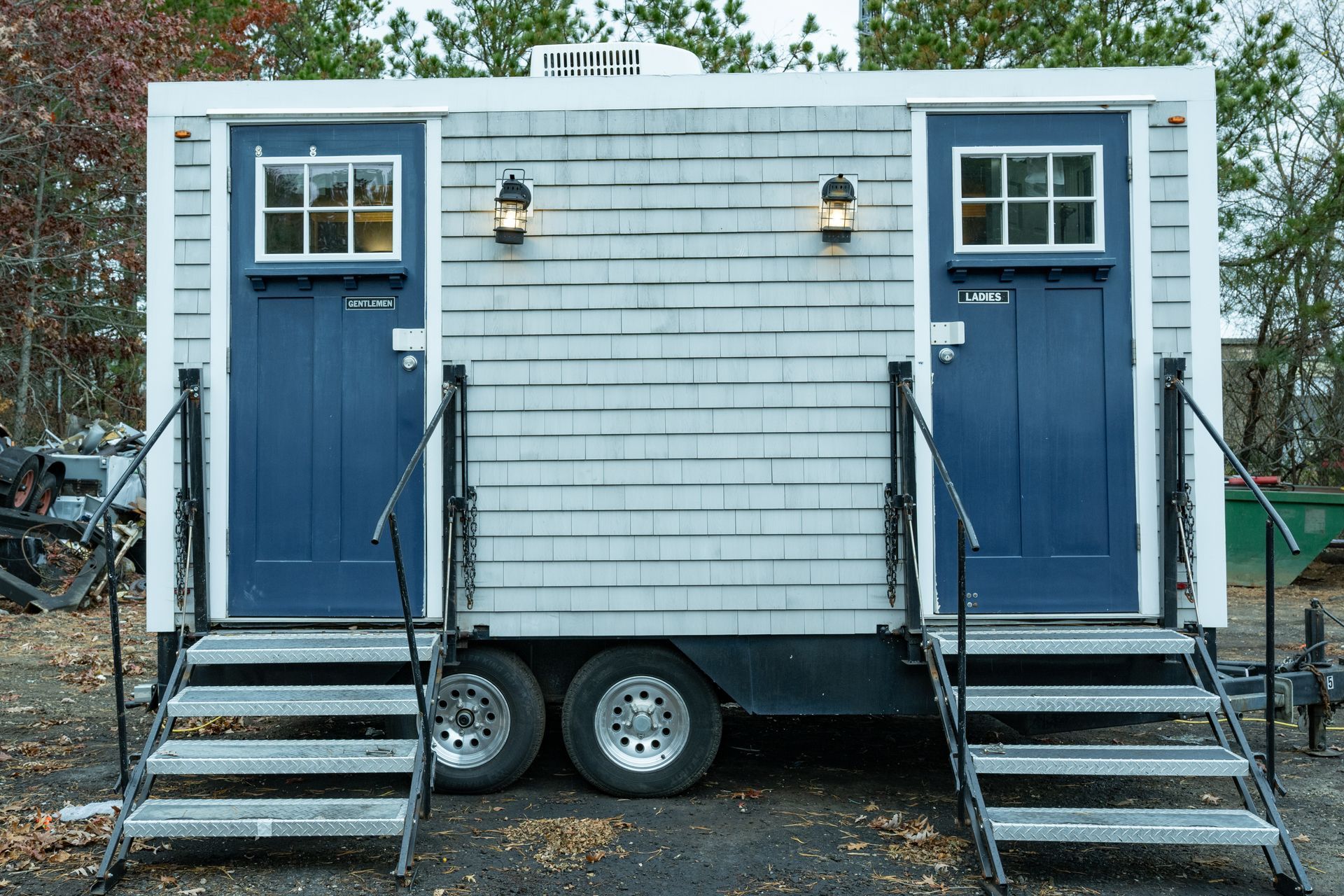 Portable restroom trailer with two blue doors, gray siding, and metal stairs.