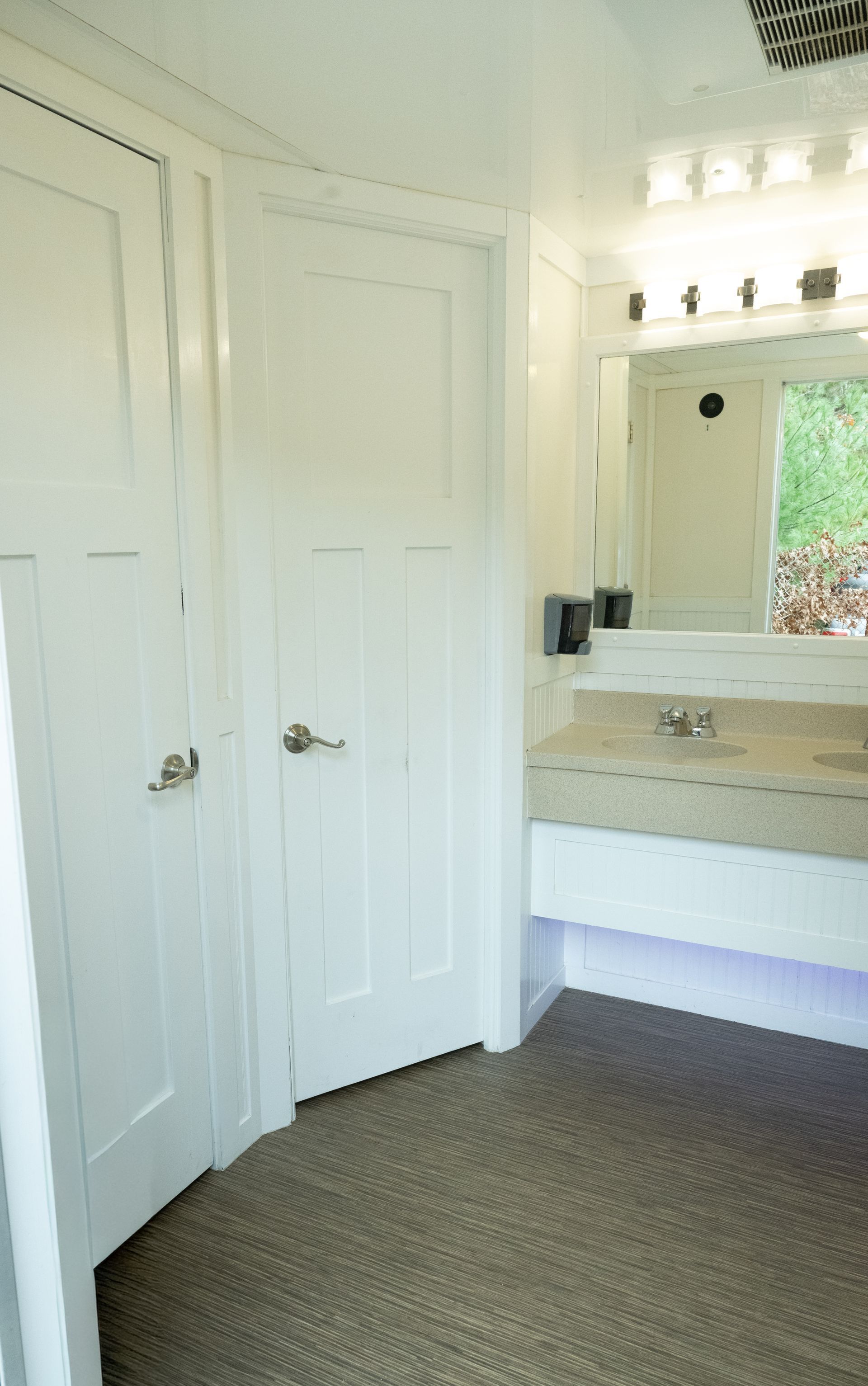 Interior of a modern restroom with two stalls, a sink, and a mirror. White walls and doors, carpeted floor.