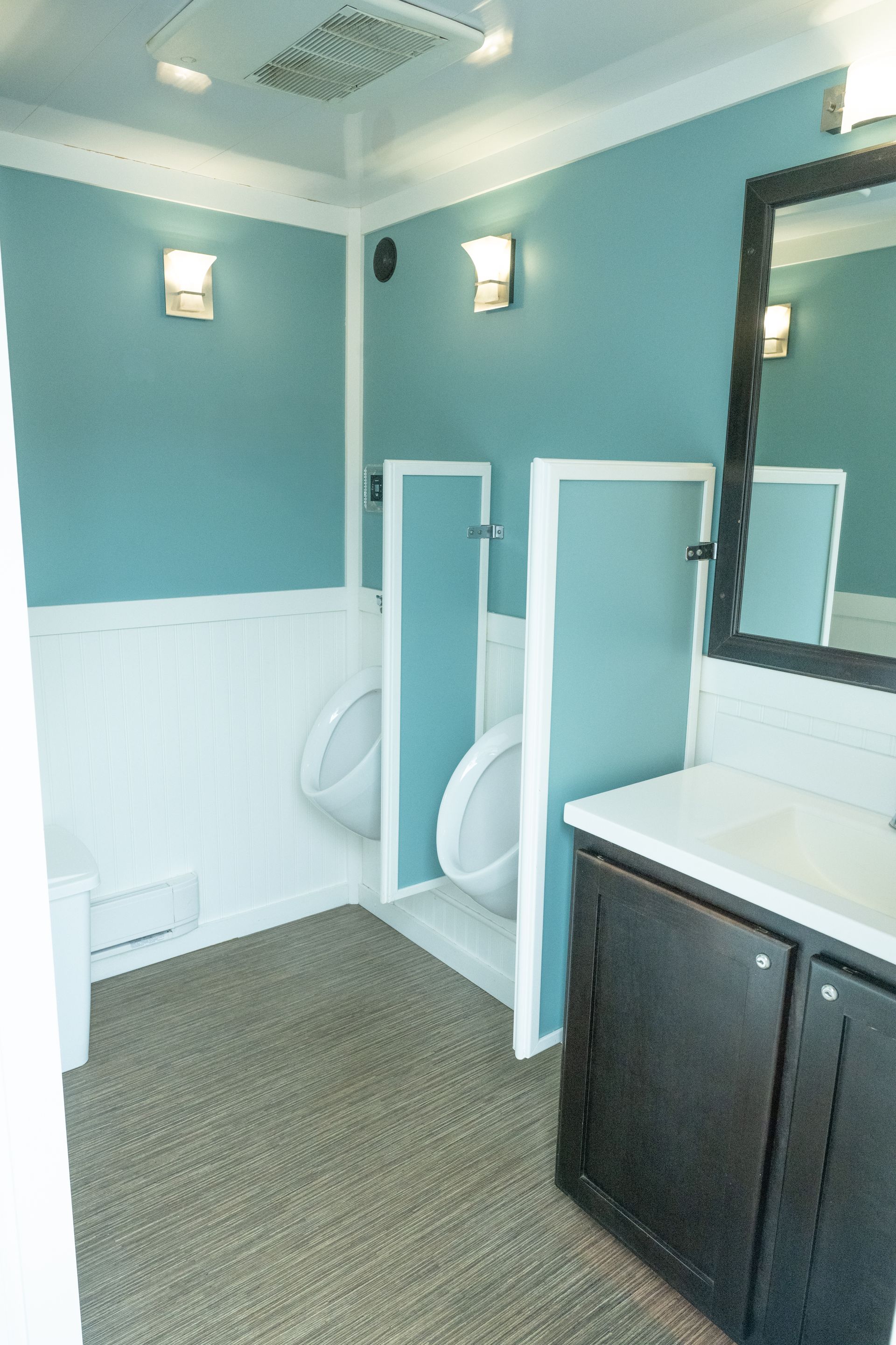 Interior of a mobile restroom with two urinals, teal and white walls, dark cabinets, and carpet flooring.