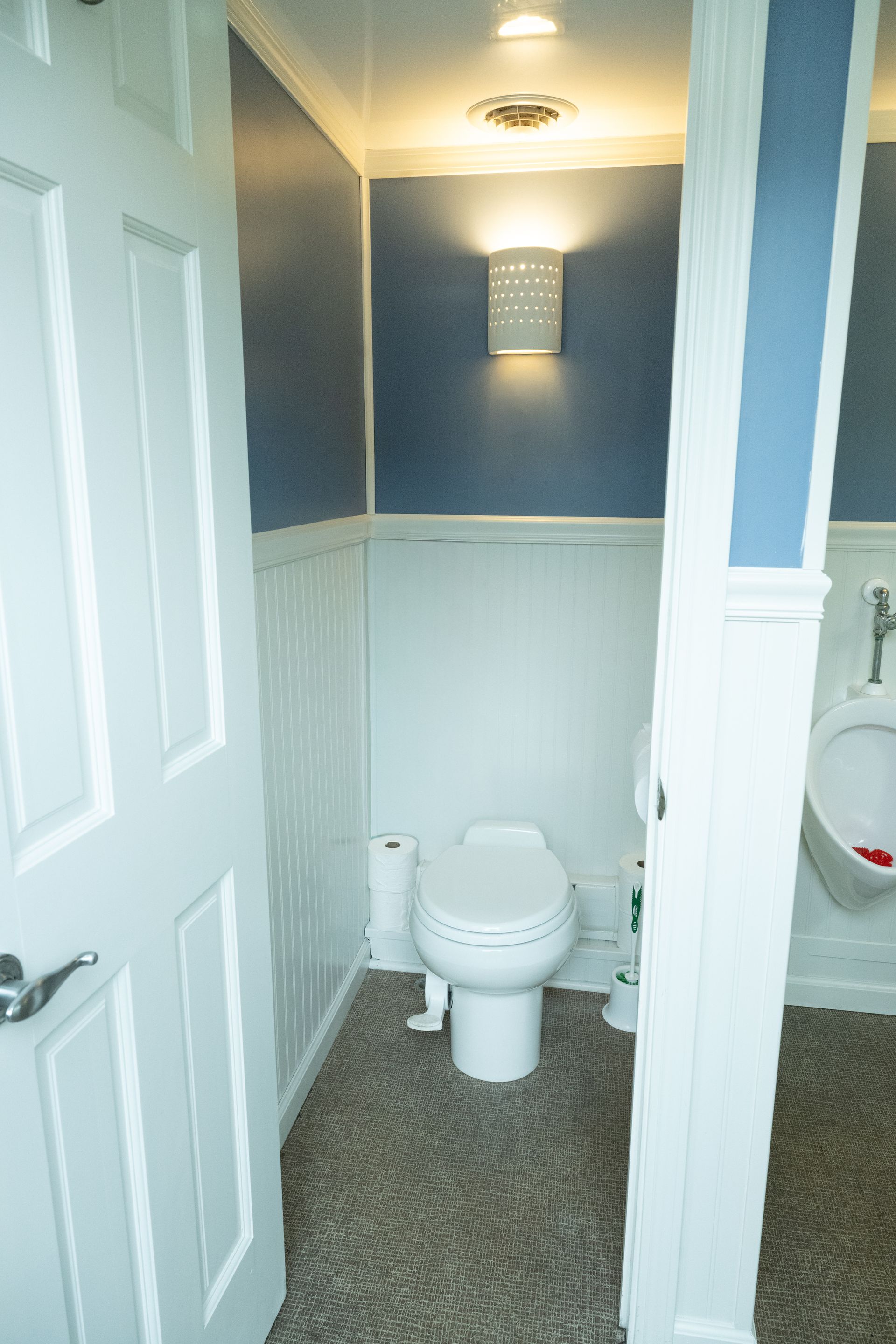 Interior of a portable restroom with a toilet and urinal. White walls and door, blue accent wall, and stone-like floor.