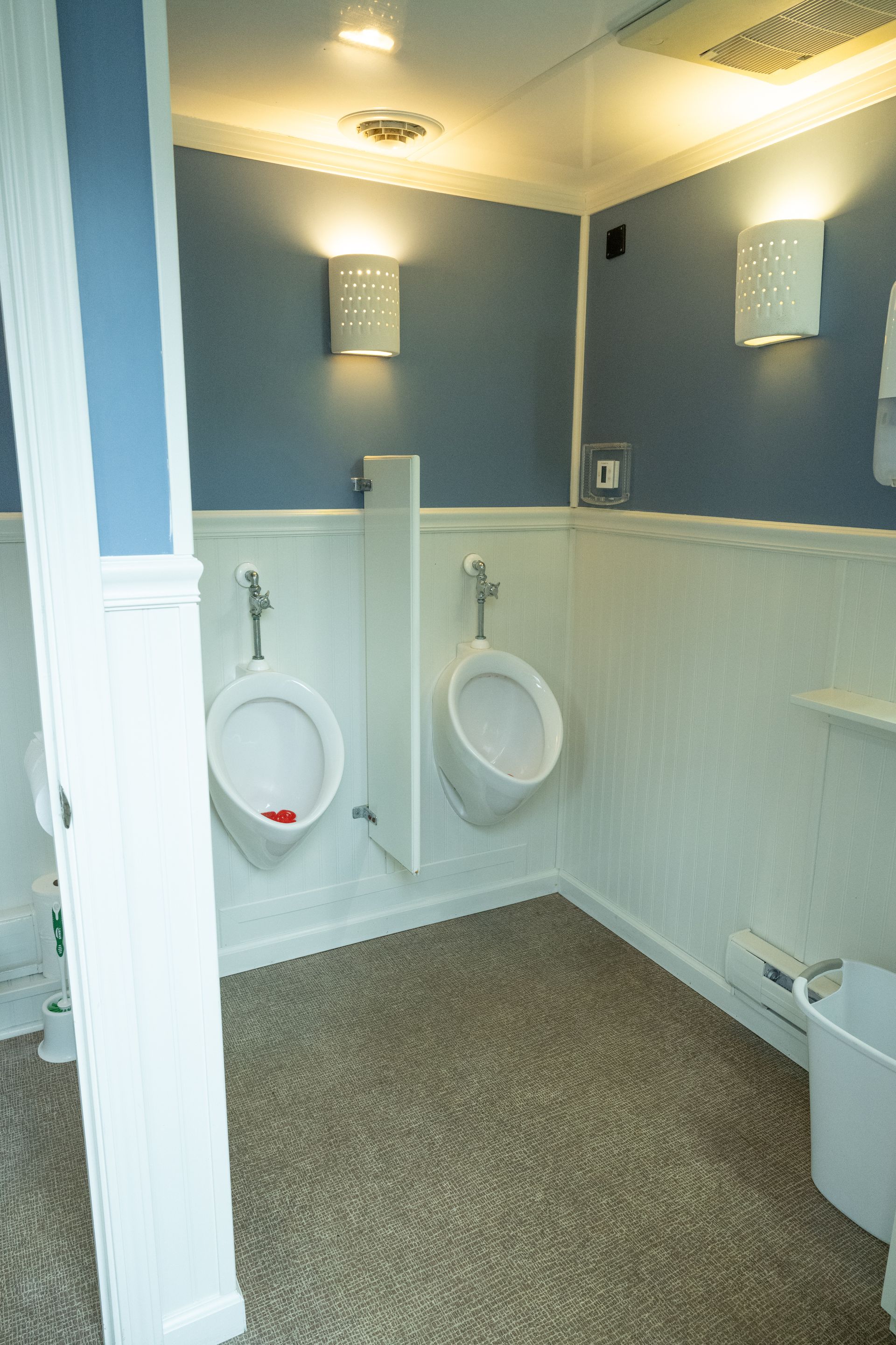 Public restroom with two urinals, blue and white walls, two wall sconces, and a carpeted floor.