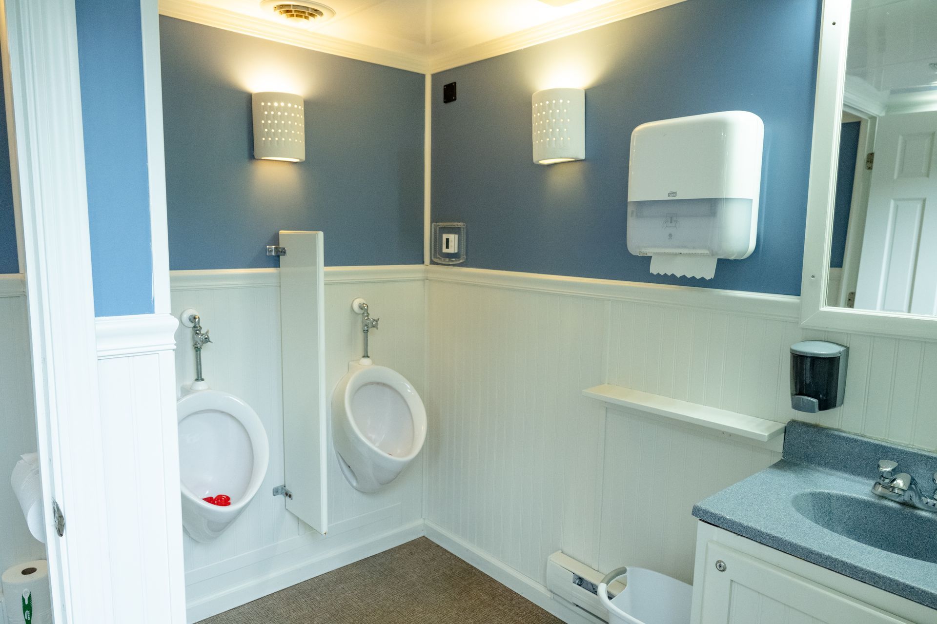 Public restroom with two urinals, a sink, and paper towel dispenser. Blue and white walls, sconce lights.