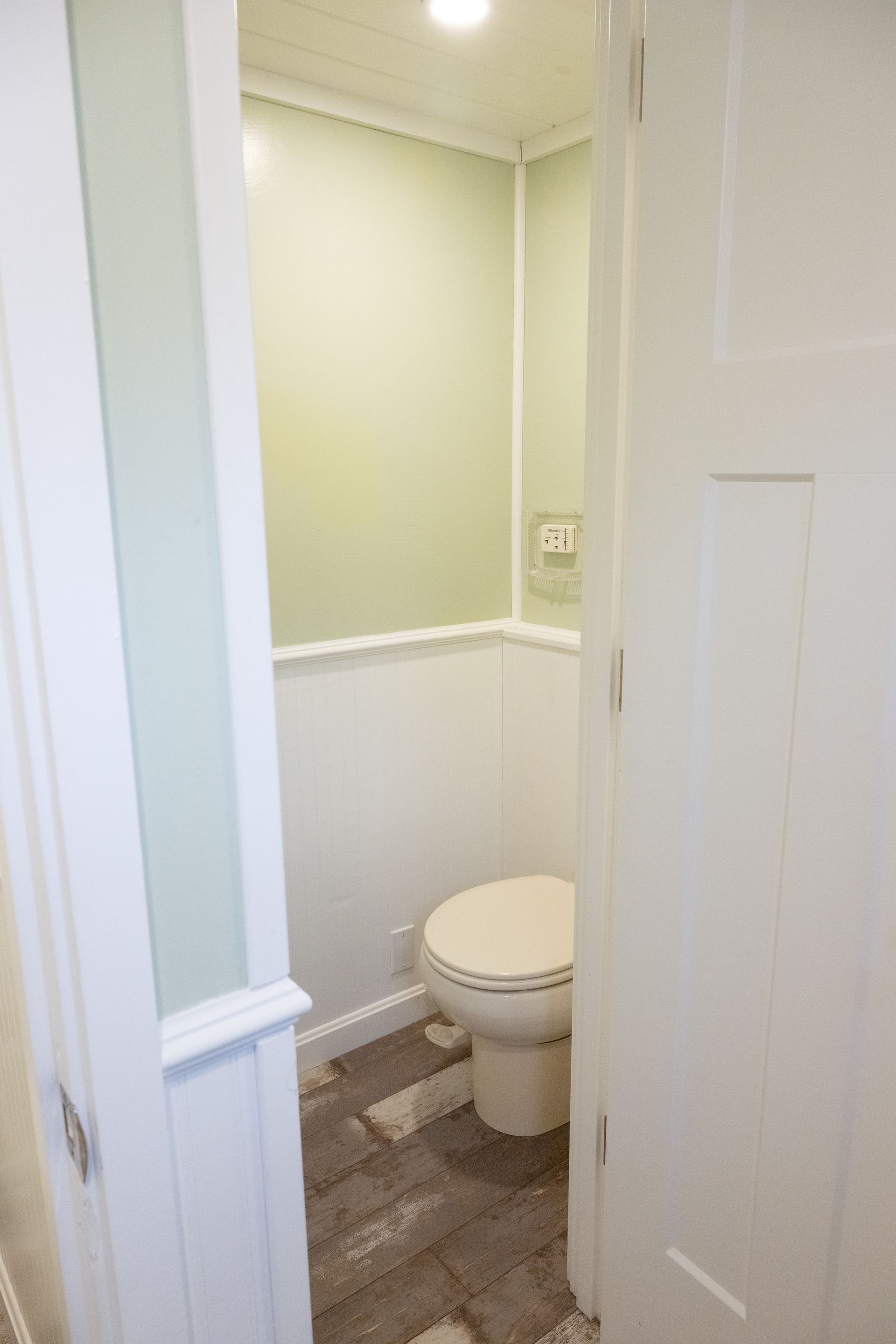 Small bathroom with white wainscoting, pale green upper walls, and wooden-look flooring; a toilet is centered.