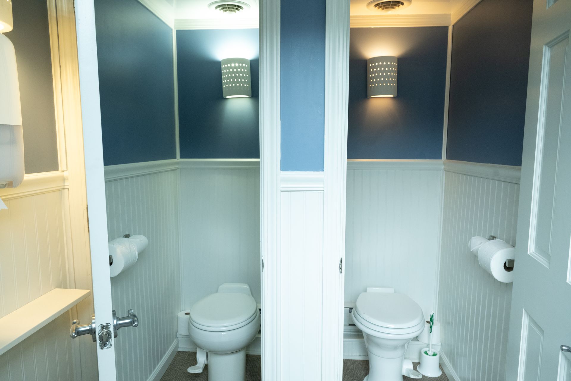 Two identical public restroom stalls with blue and white walls and sconce lighting.