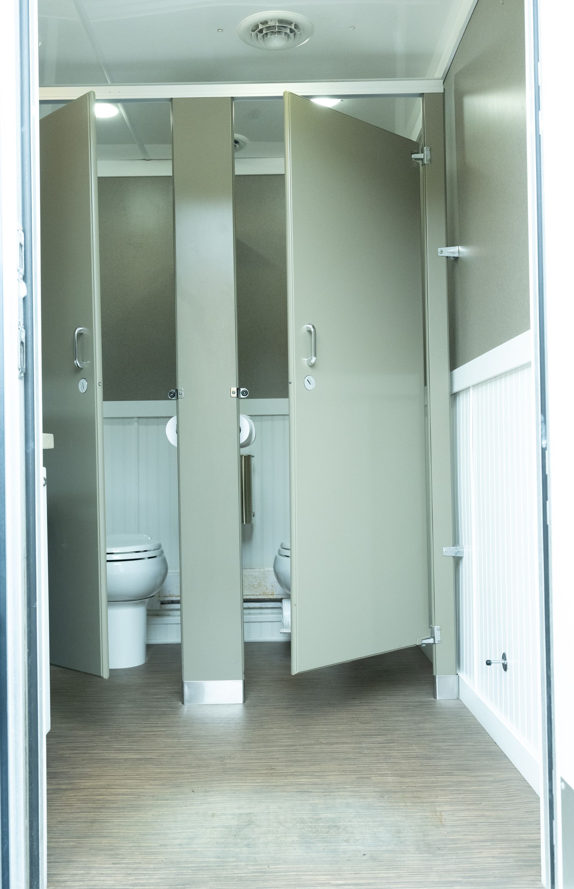 Interior of a mobile restroom trailer with three stalls. Beige doors, wood-look floor, and white wainscoting.