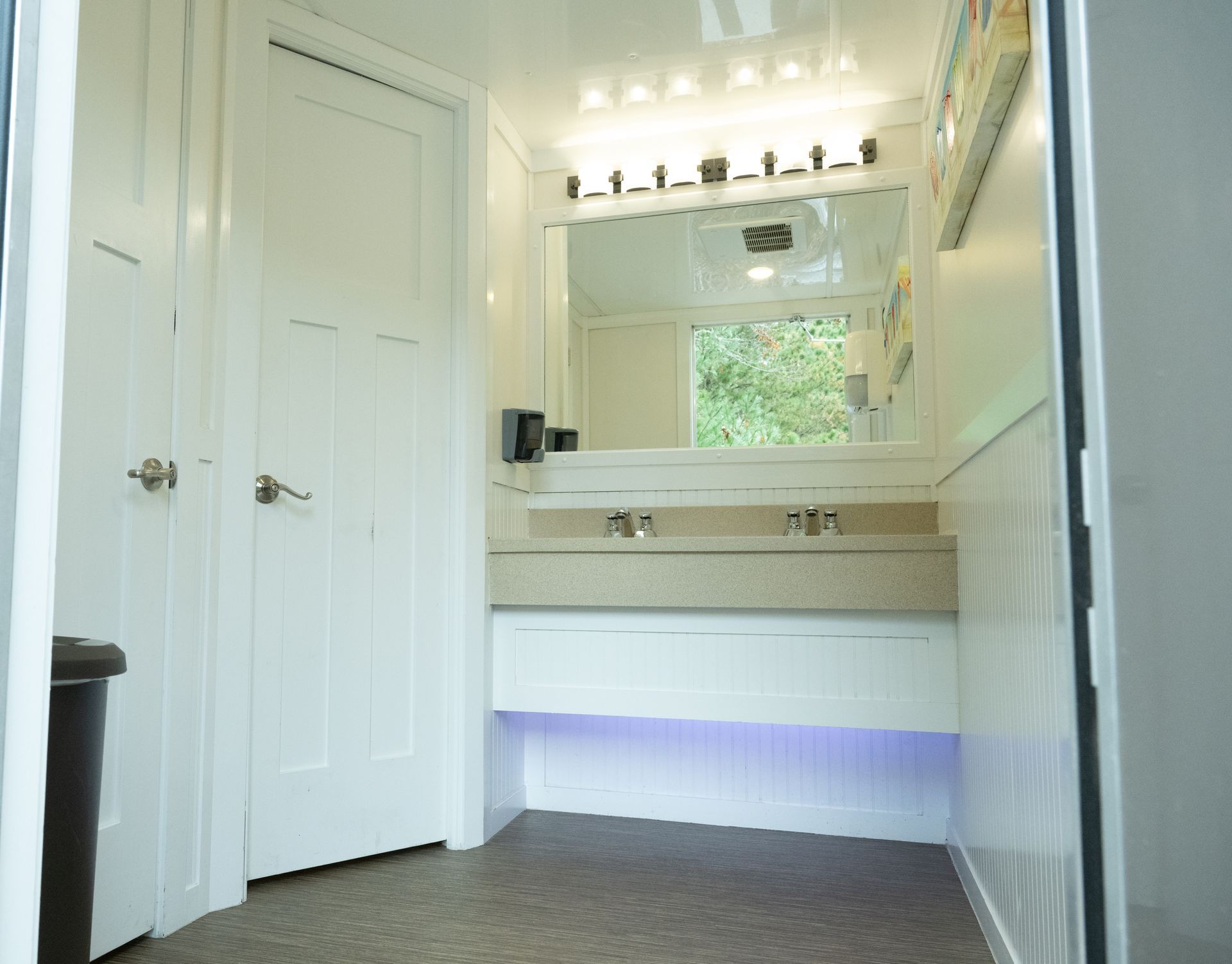 Interior view of a portable restroom with white walls, a sink area, and a closed door.