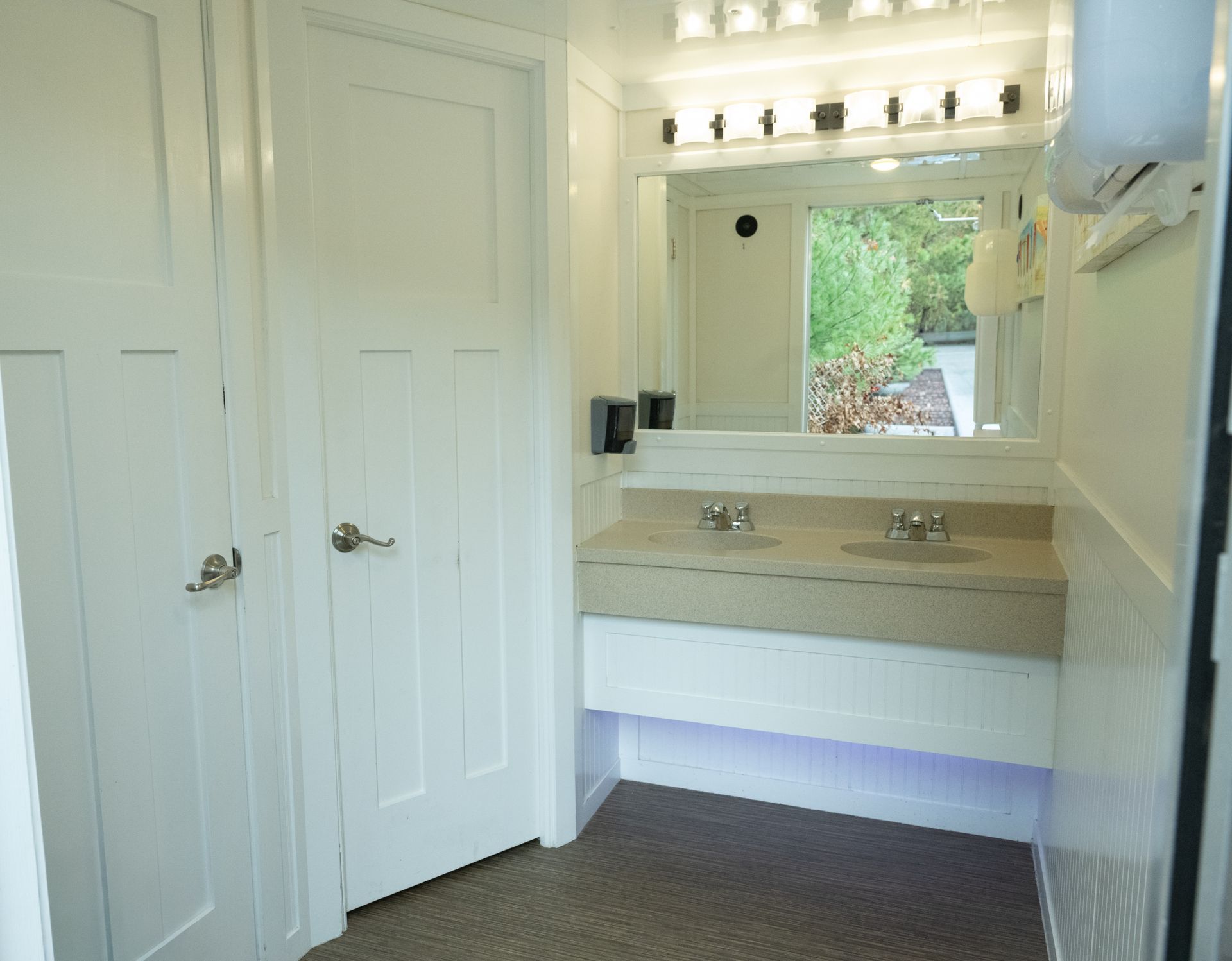 Interior of a clean, white portable restroom with a double sink vanity and doors.