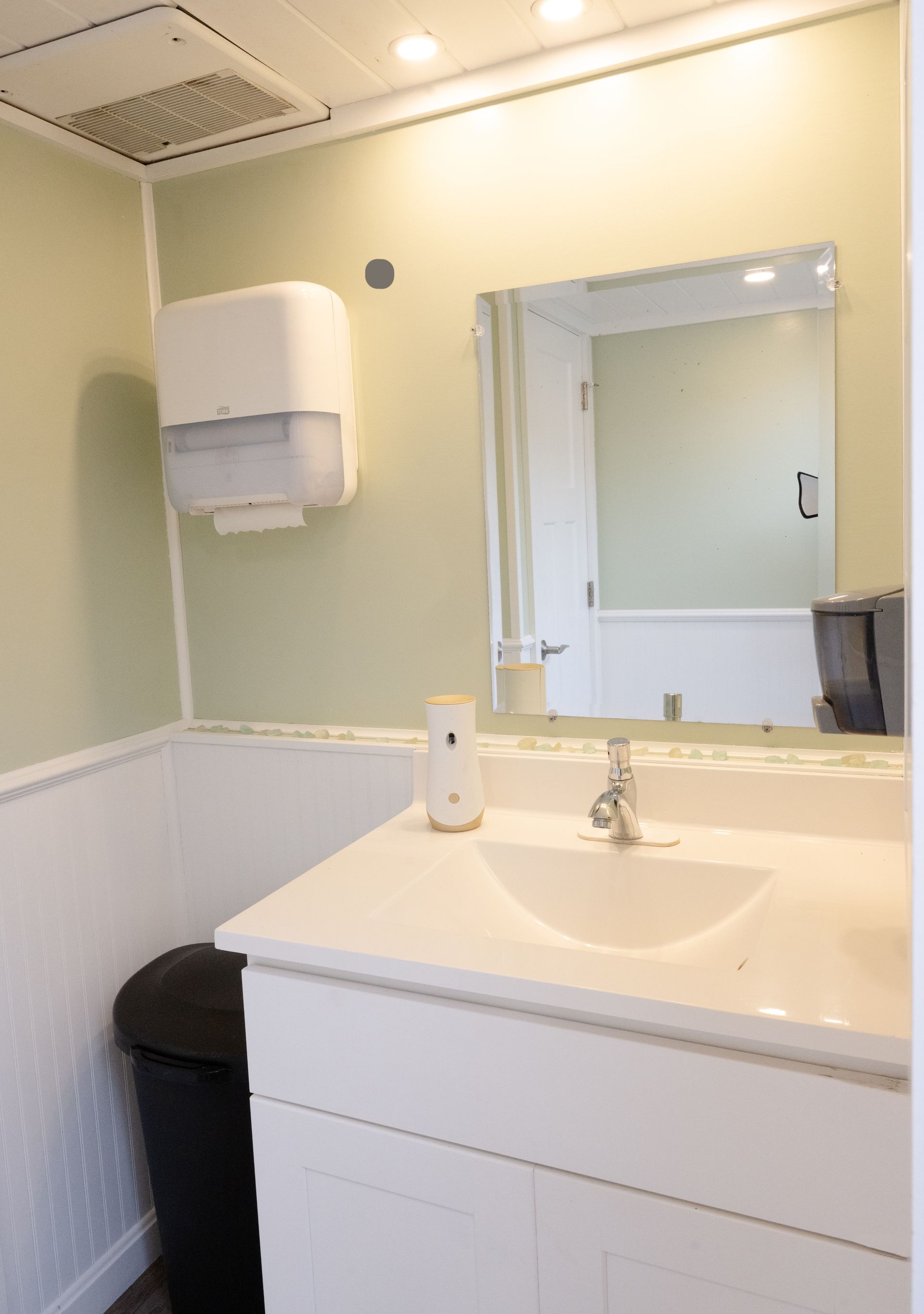 Bathroom interior: White sink, mirror, paper towel dispenser, trash can, and light green walls.