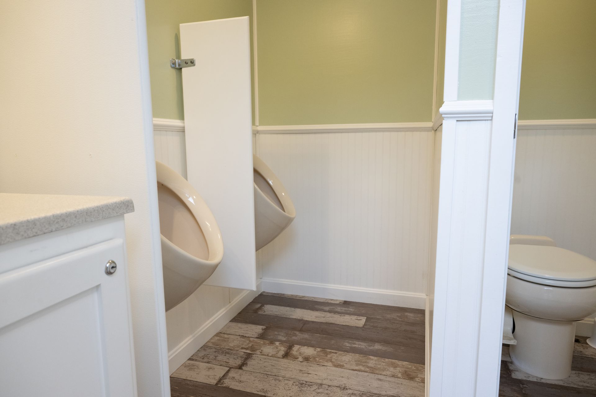Public restroom with urinals and toilet, white walls, light green upper wall, and wood-look flooring.