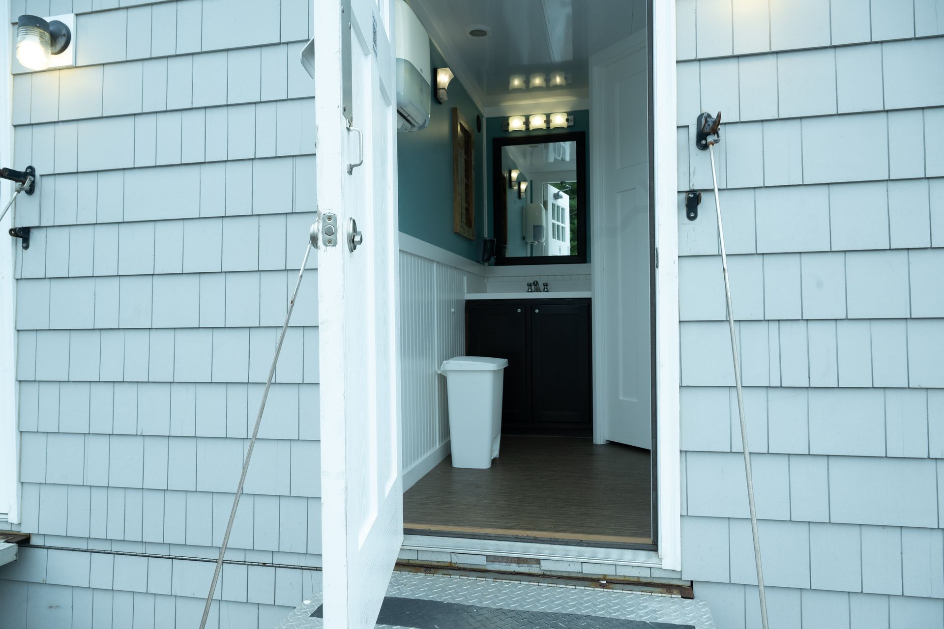 Exterior of a bathroom with an open door. Light blue shingled siding, white door, interior includes vanity, mirror and trash can.